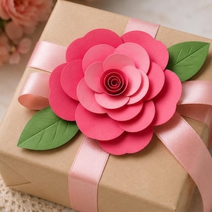 May include: A square gift box wrapped in plain brown paper, tied with a smooth light pink satin ribbon. A large, layered paper flower in shades of vibrant pink and red sits on top, accented by two pointed green paper leaves with visible veins.