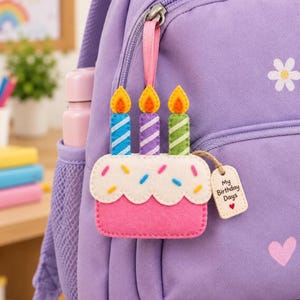 May include: A felt birthday cake keychain with three candles in blue, purple, and green, featuring yellow flames and colorful sprinkles on a white frosting top. A small wooden tag attached by a jute string displays the text My Birthday Days.