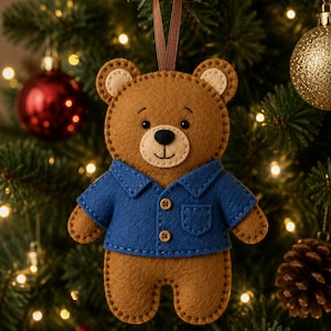 May include: A brown felt teddy bear ornament wearing a blue collared shirt with two small wooden buttons and a chest pocket. The bear has cream-coloured inner ears and a black stitched nose and mouth, suspended by a thin brown ribbon loop.