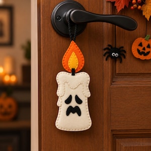 May include: A felt hanging decoration shaped like a white candle with a sad face and a yellow and orange flame. The candle is suspended by a black cord from a dark metal door handle. A small black spider and an orange pumpkin are also visible.