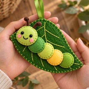 May include: A felt ornament shaped like a green leaf with a multi-colored caterpillar attached to the surface. The caterpillar has a light green head with brown antennae, pink cheeks, and a body made of green, yellow, and lime felt circles.