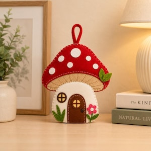 Può includere: Un ornamento in feltro a forma di fungo con cappello rosso a pois bianchi, parte inferiore beige e gambo bianco. Il gambo presenta una porta ad arco marrone, una piccola finestra circolare, dettagli in erba verde e un fiore rosa con stelo.