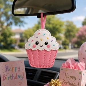 May include: A felt cupcake hanging ornament with a pink base and white frosting featuring colorful embroidered sprinkles, a smiling face with black eyes, and pink cheeks. The cupcake is suspended by a pink ribbon from a car rearview mirror.