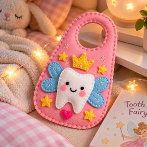May include: A pink felt door hanger featuring a white tooth character with blue wings, a yellow crown, and yellow stars. The tooth has black eyes, a black smile, and pink cheeks. A small pink heart is stitched below the tooth character.