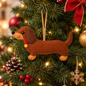 May include: A brown felt dachshund ornament with a tan face, dark brown ears, a red collar with a yellow tag, and a black nose. The dog-shaped decoration features visible stitching along the edges and hangs from a tan string loop.