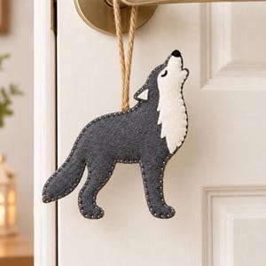 Puede incluir: Un adorno de fieltro con forma de lobo en gris oscuro y blanco, representado en posición de aullido con la cabeza hacia arriba. La figura presenta costuras claras visibles en los bordes y está colgada de un cordón trenzado de color beige.