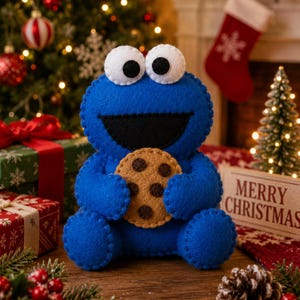 May include: A blue felt plush character with large white eyes and black pupils, holding a tan felt cookie with dark brown chocolate chips. The character has a wide black mouth and is constructed with visible blanket stitching along the edges.