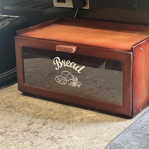 May include: A dark wooden bread bin with a glass front. The bin is a deep reddish-brown with a lighter brown lid. The word "Bread" is written in a cursive font on the glass, along with a bread graphic.