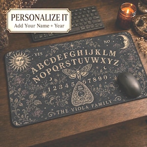 May include: A rectangular desk mat featuring a dark, ornate spirit board design with the alphabet, numbers 0-9, a sun, a moon, and a moth. The text reads "YES", "NO", "EST. 2026", and "THE VIOLA FAMILY". It includes a planchette graphic.