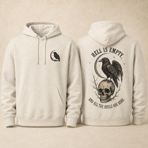 May include: A heathered beige hooded sweatshirt featuring a small black raven graphic on the front left chest. The back displays a large illustration of a raven perched on a skull beneath a crescent moon with the text HELL IS EMPTY AND ALL THE DEVILS ARE HERE.