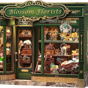 Puede incluir: Una tienda de flores en miniatura de Blossom Florists con fachada verde y adornos dorados. La tienda presenta un letrero con el nombre de la tienda, exhibiciones de flores y un pequeño carro. La tienda está decorada con flores y tiene un interior detallado.
