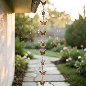 May include: A decorative rain chain with butterfly-shaped links. The butterflies have a brown and clear design, hanging from a gutter in a garden setting. The rain chain is suspended in the rain, with a blurred background of greenery and a stone pathway.