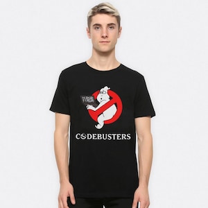 以下が含まれることがあります： 黒いTシャツ。白いお化けのようなキャラクターがメガネをかけ、ノートパソコンを持っています。赤い禁止マークの中にいます。「CODEBUSTERS」の文字が下にプリントされています。半袖Tシャツです。
