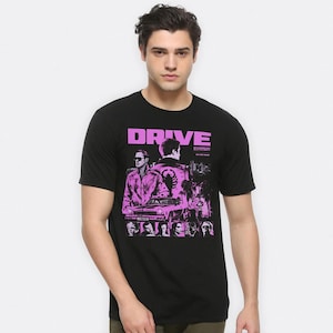 May include: Black t-shirt featuring the word "DRIVE" in pink block letters. The shirt has a graphic of a movie poster with actors and a car in pink and black. The shirt is made of a soft material.