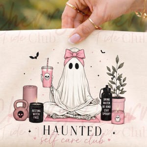 Op de afbeelding: Een lichtbeige draagtas met een grillige illustratie. Een spook met een roze strik zit in een yogahouding en houdt een drankje vast. De tas heeft de tekst "HAUNTED self care club" en andere griezelige items.