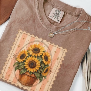Puede incluir: Una camiseta marrón de manga corta con un gráfico de girasoles en una maceta dentro de un marco rectangular con rayas color melocotón y estrellas. Un collar de cadena plateada descansa en el cuello. La etiqueta dice Comfort Colors.