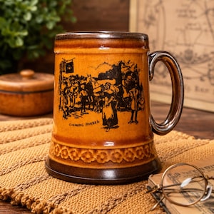 Może przedstawiać: Cylindryczny ceramiczny kufel z błyszczącym bursztynowo-brązowym szkliwem. Na zewnątrz znajduje się czarna ilustracja przedstawiająca ludzi i konie z napisem "CHANGING HORSES". Podstawa posiada ozdobny wytłaczany pas z motywem kwiatowym.