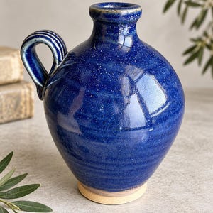 Puede incluir: Una jarra de cerámica azul cobalto con un acabado de esmalte brillante y moteado. El recipiente presenta un cuerpo redondeado, un cuello estrecho con borde acampanado y un asa única con tres crestas verticales. La base no tiene esmalte.