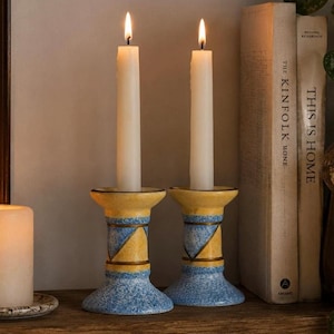 Può includere: Due portacandele in ceramica con un motivo maculato giallo e blu e accenti geometrici triangolari. Ogni portacandele sostiene una candela affusolata color bianco sporco, alta e sottile, con una fiamma accesa. Il design include bande.
