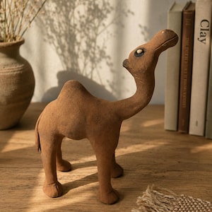 Puede incluir: Una figura de camello marrón con una superficie texturizada sobre una mesa de madera. El camello tiene un cuello largo y mira hacia arriba. Un jarrón con flores secas y una pila de libros están en el fondo.