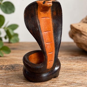 May include: A hand-carved wooden cobra figurine featuring a dark brown exterior and a lighter orange-brown interior on the hood and body. The snake is coiled at the base, with its hood flared upward and a small black dot representing the eye.