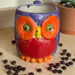May include: A ceramic owl-shaped mug with a purple body, orange eyes, and a red beak. The mug has a handle and is filled with coffee beans. The mug is sitting on a wooden surface.