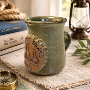 Könnte beinhalten: Ein salbeigrüner Keramikbecher mit einem strukturierten, kreisförmigen Tonmedaillon auf der Vorderseite. Das Medaillon zeigt ein Segelboot-Relief und den geprägten Text "SIR FRANCIS DRAKE" sowie "DMC" am unteren Rand. Der Becher hat eine glatte, gesprenkelte Oberfläche.