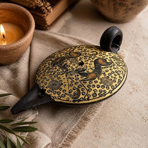 May include: An antique, black and gold oil lamp with a long spout and a handle. The lamp features a detailed design of figures and floral patterns. The lamp is set on a beige cloth, with a lit candle and other pottery in the background.