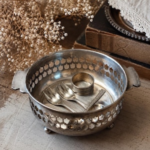 Pode incluir: Uma taça redonda de metal prateado com um padrão decorativo de treliça nas laterais, duas pequenas alças laterais e três pés em forma de esfera. Dentro da taça estão dois pequenos garfos prateados e um anel de guardanapo liso prateado.