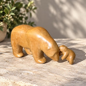 Puede incluir: Una escultura de madera tallada, lisa y de color marrón claro, que representa a un oso adulto grande junto a un osezno. Las figuras tienen un acabado pulido con vetas de madera visibles, mostrando al oso adulto inclinado hacia el osezno.