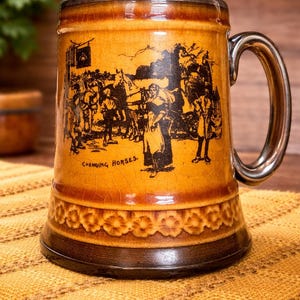 Könnte beinhalten: Vintage Bierkrug aus Keramik mit einer detaillierten schwarz-braunen Illustration, die eine Szene mit Pferden und Menschen darstellt, mit dem Text "Changing Horses". Der Krug hat einen silberfarbenen Henkel und Rand sowie ein dekoratives Blumenmuster.