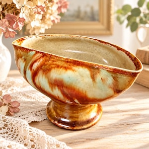 May include: A ceramic pedestal bowl with an oval, slanted rim. The exterior and interior feature a glossy glaze with swirling patterns of deep brown, burnt orange, and soft mint green over a cream-colored base, set on a circular foot.