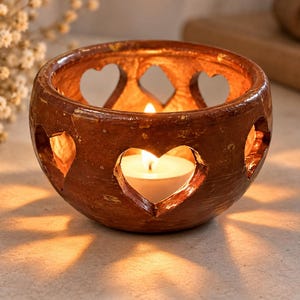 Puede incluir: Un portavelas redondo de cerámica marrón con acabado brillante y múltiples recortes en forma de corazón en los laterales. Una pequeña vela de té blanca se encuentra en el interior, emitiendo una luz cálida que proyecta sombras.