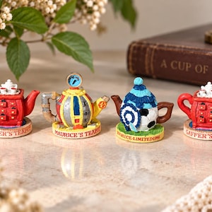 Pode incluir: Quatro bules decorativos em miniatura dispostos em fila. Dois bules vermelhos rotulados como Gaffer's Teapot, um bule amarelo e cinza rotulado como Maurice's Teapot e um bule com tema de futebol rotulado como Bruce Limited 1996 com um gorro azul e branco.