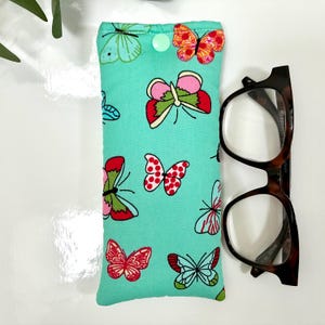 May include: A teal glasses case with a colorful butterfly pattern and a green button closure. Tortoiseshell eyeglasses rest beside the case. The butterflies are red, pink, orange, and blue.