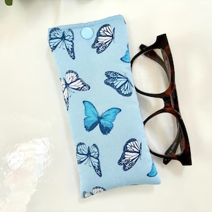 May include: A light blue glasses case with a pattern of blue and white butterflies. The case is rectangular with a button closure. Next to the case are brown tortoiseshell eyeglasses.
