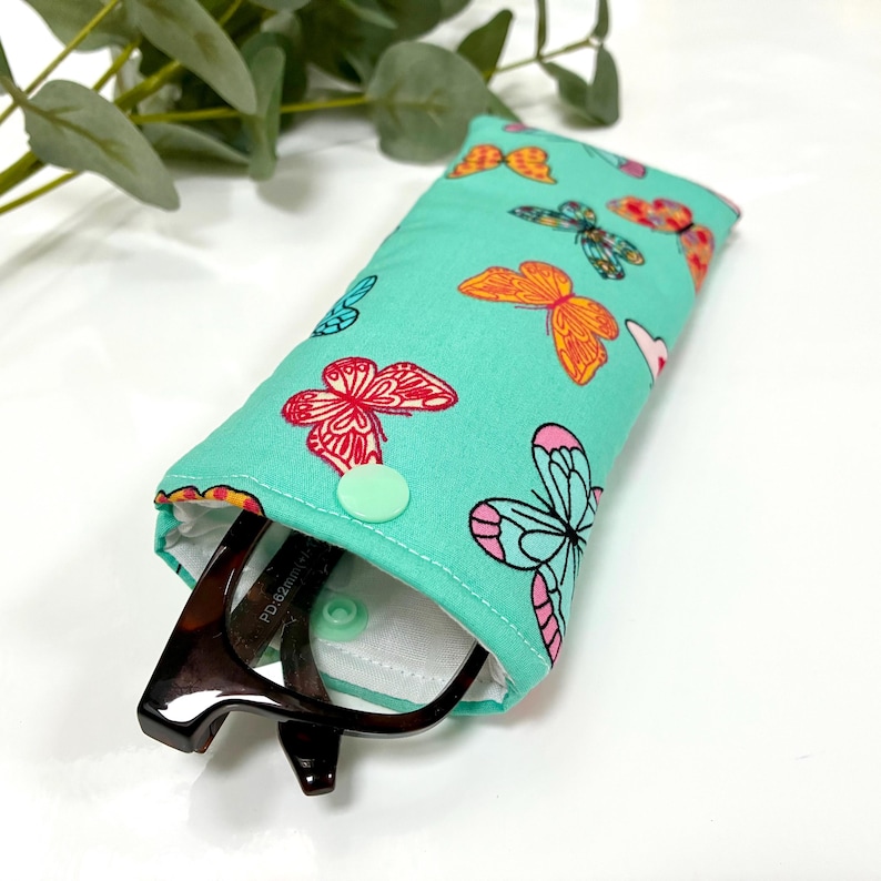 May include: A teal glasses case with a colorful butterfly pattern. The case is open, revealing a pair of eyeglasses. The butterflies are in shades of pink, orange, and blue. A mint green snap closure secures the case.