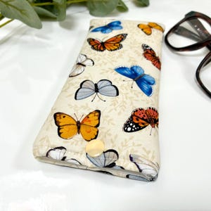 May include: A beige eyeglasses case adorned with colorful butterfly patterns. The butterflies are in shades of orange, blue, yellow, and white, set against a backdrop of subtle leaf designs. The case has a snap closure and is designed to protect eyewear.