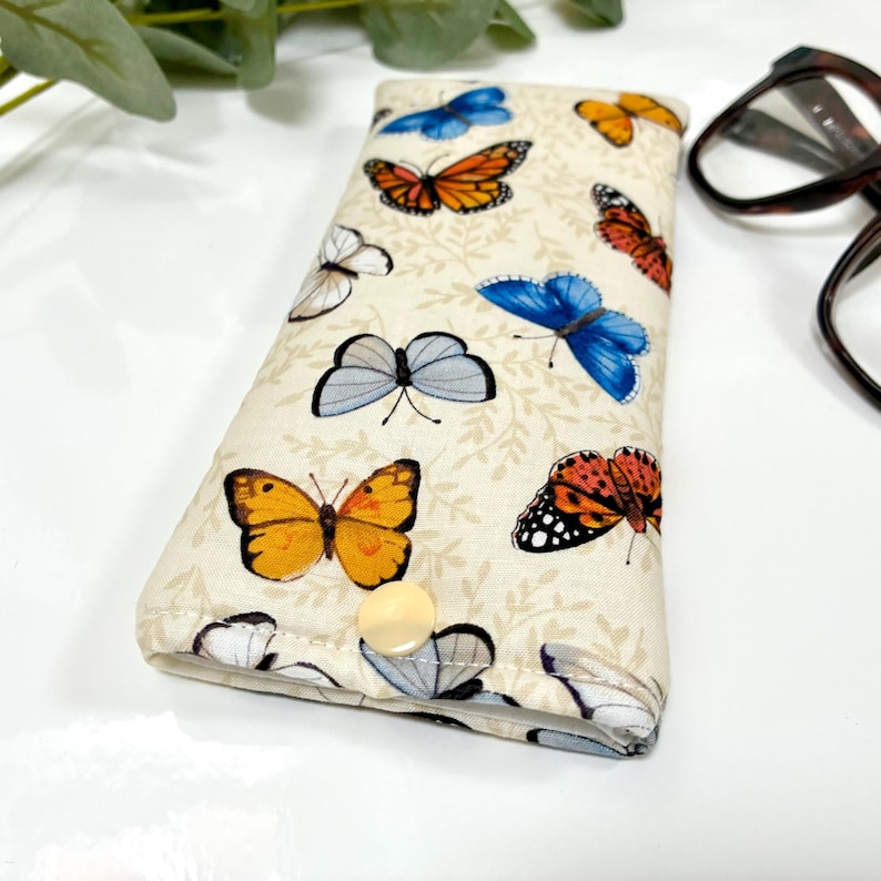 May include: A beige eyeglasses case adorned with colorful butterfly patterns. The butterflies are in shades of orange, blue, yellow, and white, set against a backdrop of subtle leaf designs. The case has a snap closure and is designed to protect eyewear.