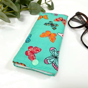 May include: A teal glasses case with a colorful butterfly pattern. The butterflies are in shades of orange, pink, red, and blue. The case has a button closure and a white interior. Eyeglasses are visible in the background.