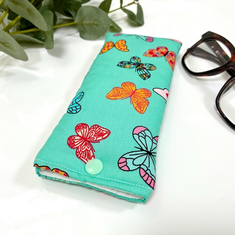 May include: A teal glasses case with a colorful butterfly pattern. The butterflies are in shades of orange, pink, red, and blue. The case has a button closure and a white interior. Eyeglasses are visible in the background.
