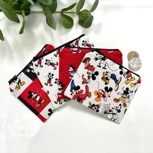 May include: Three small zippered pouches made of fabric featuring a repeating pattern of Mickey Mouse in various poses and outfits. The pouches are coloured in red, white, and black, with one pouch featuring a red background and two with white.