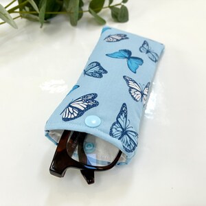 May include: A light blue glasses case with a pattern of blue and white butterflies. The case is open, revealing a pair of eyeglasses. The case is closed with a light blue button.