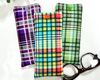 May include: Three plaid eyeglass cases in purple, teal, and green, each with a button closure. A pair of brown-framed glasses rests nearby. The cases feature a variety of colorful plaid patterns, offering a stylish way to protect eyewear.