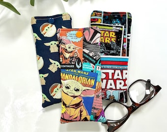 May include: Three rectangular fabric pouches with snap closures. One features a dark blue pattern of small green creatures, another displays a colorful Star Wars comic book print, and the third shows a Star Wars collage. Brown tortoise shell glasses.