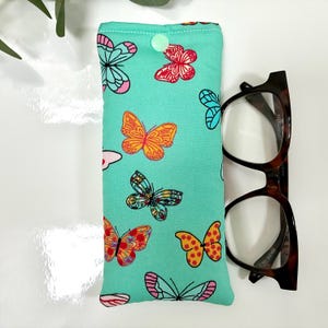 May include: A teal glasses case with a button closure, adorned with colorful butterfly patterns. Next to the case are brown tortoiseshell eyeglasses.