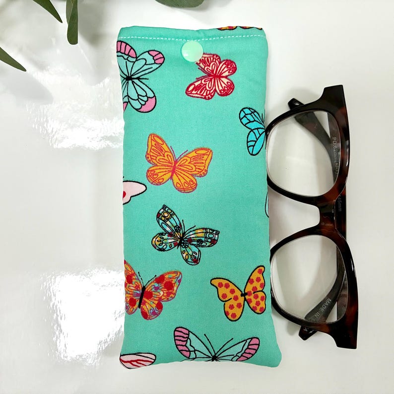 May include: A teal glasses case with a button closure, adorned with colorful butterfly patterns. Next to the case are brown tortoiseshell eyeglasses.