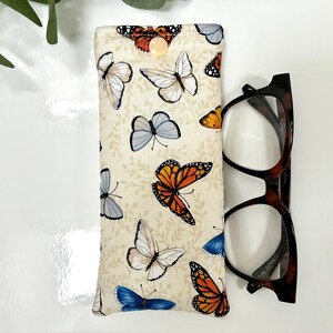 May include: A beige glasses case with a butterfly pattern, featuring orange, blue, and white butterflies. The case has a button closure and is next to a pair of tortoiseshell eyeglasses.