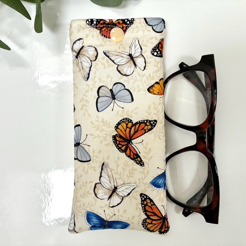 May include: A beige glasses case with a butterfly pattern, featuring orange, blue, and white butterflies. The case has a button closure and is next to a pair of tortoiseshell eyeglasses.