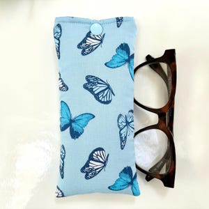 May include: A light blue glasses case with a pattern of blue and white butterflies. The case is closed with a light blue button. A pair of brown tortoiseshell glasses is next to the case.